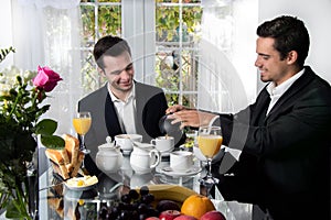 Friends or Gay couple sitting at table in front of patio doors eating breakfast and smiling