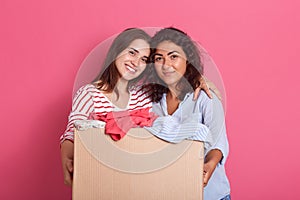 Friends girls with happy facial expression looking directly at camera while hugging, holding box full of clothes for secondary