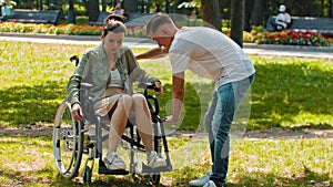 Friends in park - a guy rides disabled woman in a wheelchair then stops and sits next to her on the ground