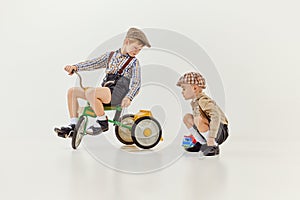 Friends playing. Boys, childred in retro clothes riding vintage bicycle over grey studio background. Concept of game