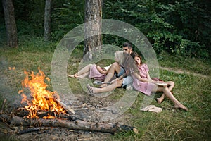 Friends relax at campfire. People at bonfire flame in green forest. Women and man at fire in camp. Camping, hiking and