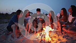 Friends sit around bonfire, drink beer, sing to guitar, fry sausages on sandy beach. Young mixed race group of men and