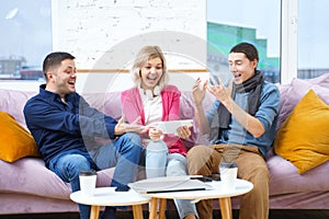 Friends sit on the couch, chatting and using a smartphone, desktop computer, laptop.