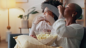 Friends on sofa with popcorn, watching tv, movie or television series together in living room. Black woman with friend