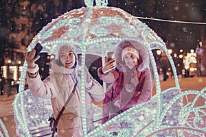 Friends taking selfie photo in illuminated fairy tale carriage with horses decorated with lights and garlands for Christmas