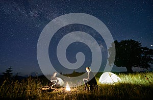 Friends travellers spending time in the mountains sitting around bonfire in camping under beautiful starry sky