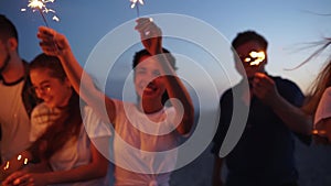 Friends walking, dancing, having fun at night party at the seaside with sparklers in their hands. Young teenagers