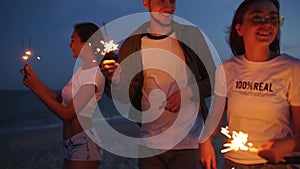 Friends walking, dancing, having fun at night party at the seaside with sparklers in their hands. Young teenagers
