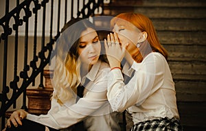 A friendship between two schoolgirls in uniform sitting on a staircase. A ginger Asian girl is whispering a secret into the ear of