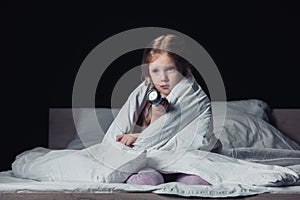Frightened child sitting under blanket and holding flashlight