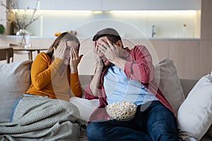 Frightened emotional young couple with popcorn watching horror film, thriller, scary movie at home