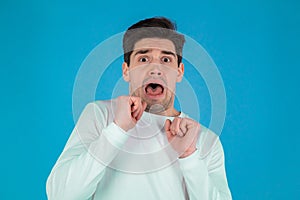 Frightened european man afraid of something and looks into camera with big eyes full of horror over blue wall background