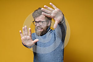 Frightened hipster in an old sweater and glasses makes a stop gesture with hands, on a yellow background.