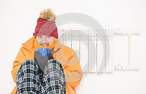 Frightened man feeling cold in hat and down jacket sitting close to radiator. Gas crisis concept