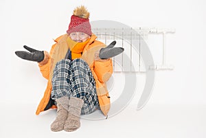 Frightened man feeling cold in hat and down jacket sitting close to radiator. Gas crisis concept