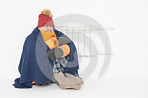 Frightened man feeling cold in hat and down jacket sitting close to radiator. Gas crisis concept