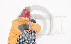 Frightened man feeling cold in hat and down jacket sitting close to radiator. Gas crisis concept