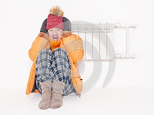 Frightened man feeling cold in hat and down jacket sitting close to radiator on white background