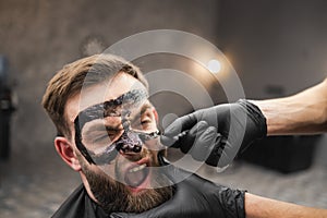 Frightened man grimaces while removing black facail skin mask in beauty salon.