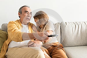 Frightened senior european spouses watching TV, man holding remote control and woman closing face, sitting on sofa