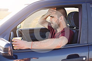 Frightened terrified dark haired unshaved man, stops his car on side of road, drinks tea, sees with shock through windshield at