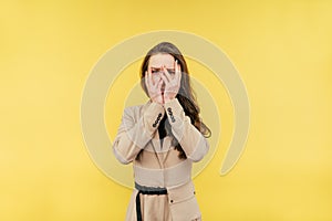 Frightened woman in a suit hid behind her hands and spied, looking at the camera on a yellow background with a sad face