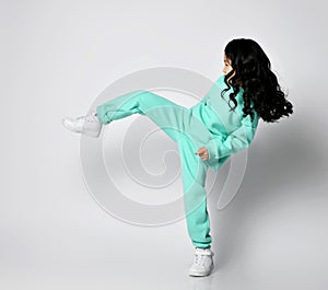 Frolic asian kid girl in pastel green, mint color hoodie and pants stands sideways kicking, playing football