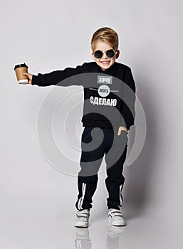 Frolic blond kid boy in black sweater with printed words holds out a cup of coffee. Translation: `Want. Can. Will Do`