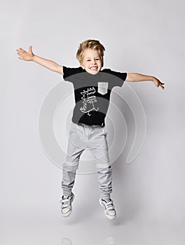Frolic blond kid boy in sunglasses, black t-shirt with dinosaur print and gray pants jumps with hands spread wide
