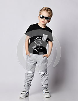 Frolic blond kid boy in sunglasses, black t-shirt with dinosaur print and gray pants stands leaning sideways