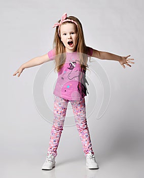 Frolic blonde kid girl in summer clothing colorful pants and t-shirt stands with her arms outstretched and screams