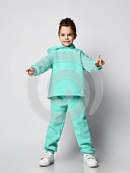 Frolic boy in modern green, mint color hoodie, pants and white sneakers stands holding hands up pointing with fingers