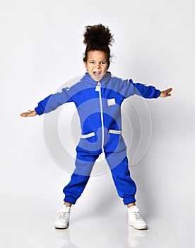 Frolic dark-skinned kid girl in blue jumpsuit stands holding arms spread wide, playing airplane and screams loudly