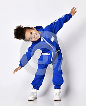 Frolic dark-skinned kid girl in blue overall jumpsuit stands leaning forward holding arms spread wide, playing airplane