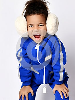 Frolic happy dark-skinned kid girl in blue overall jumpsuit and earmuffs stands leaning forward with her hands on knees