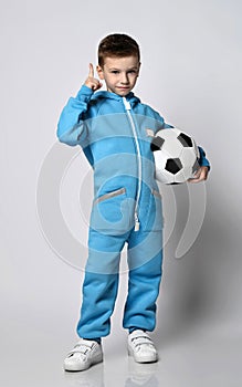 Frolic kid boy in blue jumpsuit with pockets stands holding soccer ball in hand and gesturing forefinger up