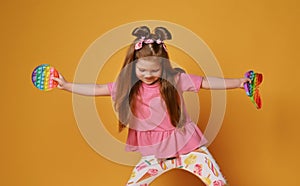 Frolic kid girl in colorful clothes holds two sensory rainbow color toys - pop it and looks down. Playing and dancing