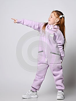 Frolic kid girl in pink modern jumpsuit stands sideways pointing with finger at upper corner, aside