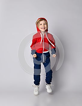 Frolic laughing blond kid boy in red and blue sport suit standing is jumping, hopping, leaping
