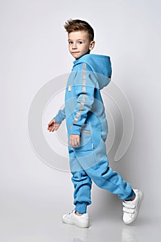 Frolic smiling kid boy in blue jumpsuit with hood and pockets with reflective stripes walks. Side view