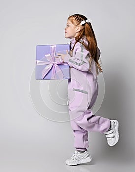 Frolic smiling kid girl in pink jumpsuit is running with big blue gift box with ribbon and bow in hands. Side view