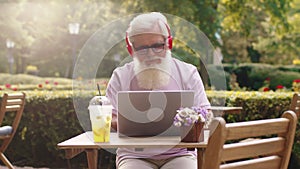 In front of the camera old man very charismatic listen music from headphones and typing something on the laptop while