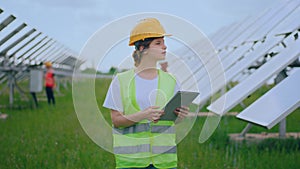 In front of the camera walking through the photovoltaic farm beautiful lady with safety helmets and equipment analysing