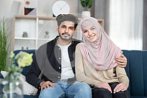 Front close up portrait of likable happy smiling young arabian couple, hugging each other, sitting on the sofa at home
