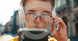 Front close-up view of the attractive smiling hipster man in eyeglasses holding the smartphone and enjoying music in his