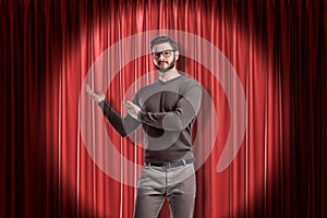 Front crop view of young handsome man in casual clothes and glasses standing in spotlight against red stage curtain and