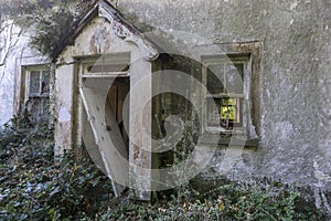 Front of derelict cottage with entrance door leaning to one side in small porch. With a broken window on either side. Vegetation o