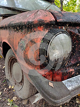 The front of a dirty old rusty car covered in rust. You can see the headlight and the wheel