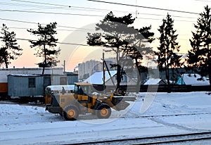 Front-end loader with wheels on a construction site in winter against the backdrop of sunset and spruce trees.