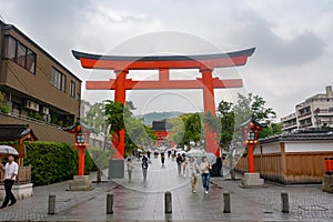 Front entrance of Fushimi Inari Shrine with large red torii gate and people walking holding umbrella
